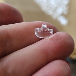 May include: A small, clear, transparent plastic button shaped like a miniature sombrero. The button features a rounded brim and a raised, conical crown, with a smooth, glossy surface that reflects light, creating a distinct, three-dimensional form.