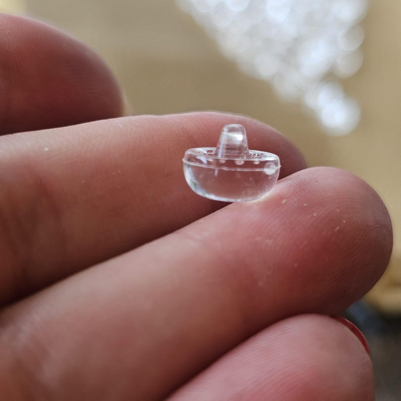 May include: A small, clear, transparent plastic button shaped like a miniature sombrero. The button features a rounded brim and a raised, conical crown, with a smooth, glossy surface that reflects light, creating a distinct, three-dimensional form.
