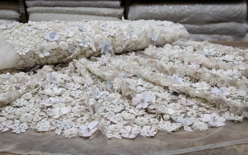 May include: White floral lace fabric with 3D flowers. The fabric is delicate and intricate, with a soft, romantic feel.