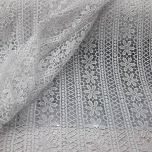 May include: White crochet lace fabric with a delicate floral pattern. The lace has a repeating pattern of small flowers and a geometric design.