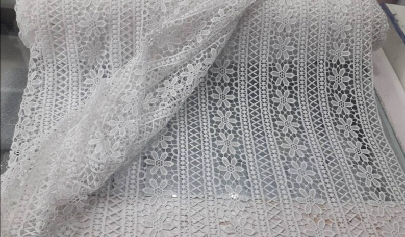 May include: White crochet lace fabric with a delicate floral pattern. The lace has a repeating pattern of small flowers and a geometric design.