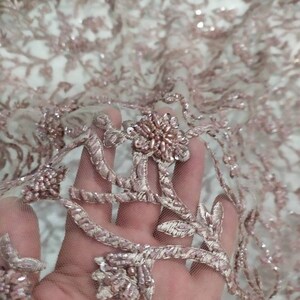 May include: Close-up of a delicate pink beaded lace fabric with floral embroidery. The fabric is sheer and has a soft, romantic feel.