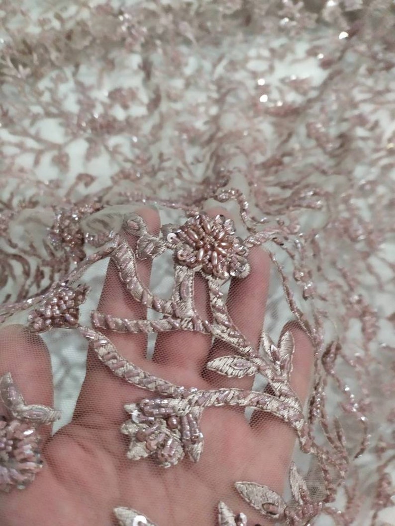 May include: Close-up of a delicate pink beaded lace fabric with floral embroidery. The fabric is sheer and has a soft, romantic feel.