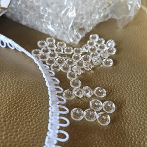 May include: A collection of small, round, clear plastic buttons scattered near a white elastic loop trim. The buttons are transparent with a smooth, circular surface and a central hole, while the trim features a continuous row of white loops.