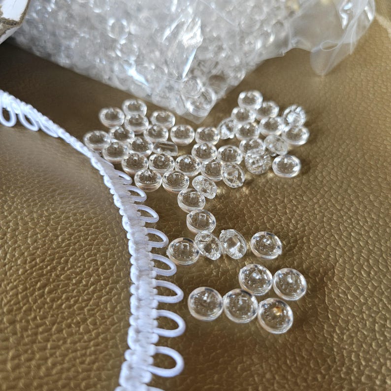 May include: A collection of small, round, clear plastic buttons scattered near a white elastic loop trim. The buttons are transparent with a smooth, circular surface and a central hole, while the trim features a continuous row of white loops.