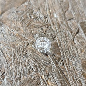 May include: A small, round, clear button featuring a faceted edge design and a central recessed area with two holes for attachment. The button is constructed from a transparent material that reflects light, creating a bright and polished appearance.