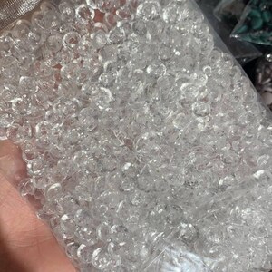 May include: A clear plastic resealable bag filled with a large quantity of small, round, transparent, faceted acrylic beads. Each bead features a multi-faceted surface that reflects light, creating a sparkling appearance throughout the collection.