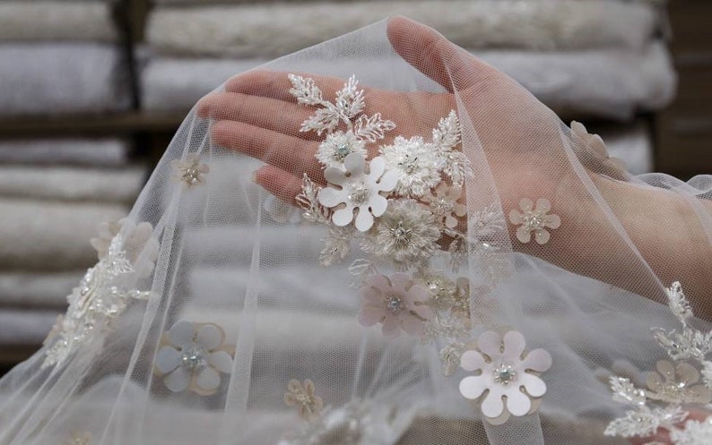 May include: White tulle fabric with delicate floral embroidery. The fabric features white and pink flowers with pearl accents.