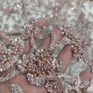 May include: Close-up of a delicate, blush-coloured fabric with intricate floral embroidery. The fabric is adorned with small, iridescent sequins and pearl-like beads, creating a luxurious and elegant look.