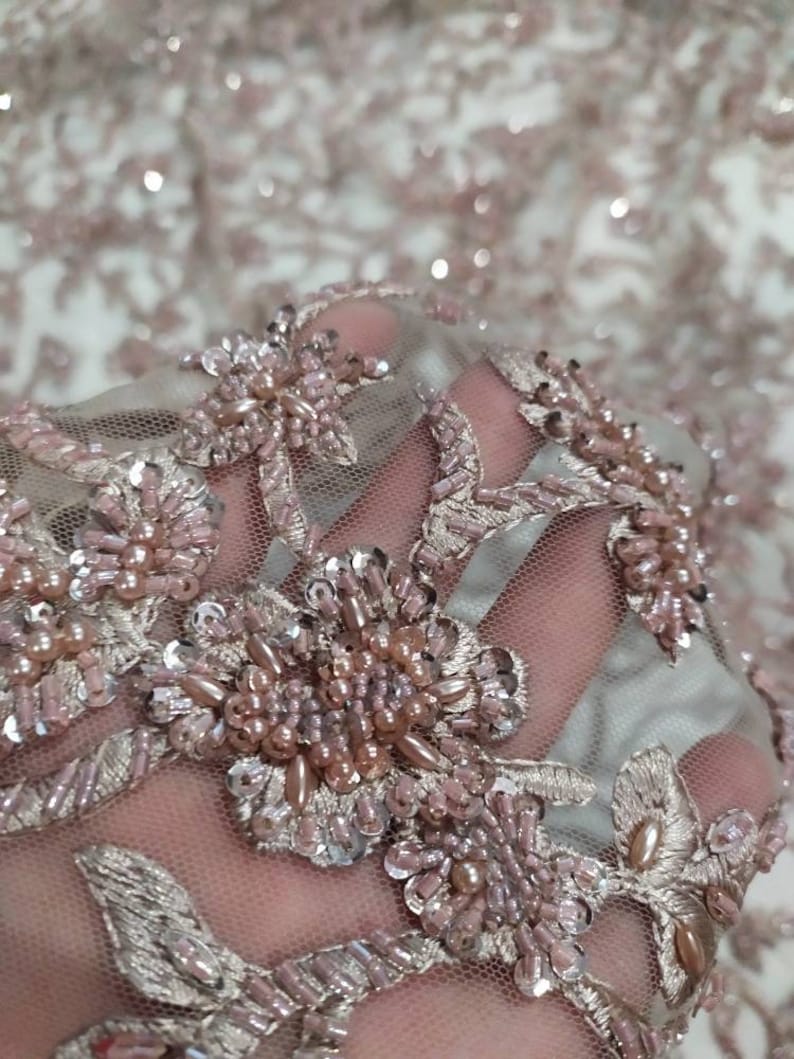 May include: Close-up of a delicate, blush-coloured fabric with intricate floral embroidery. The fabric is adorned with small, iridescent sequins and pearl-like beads, creating a luxurious and elegant look.