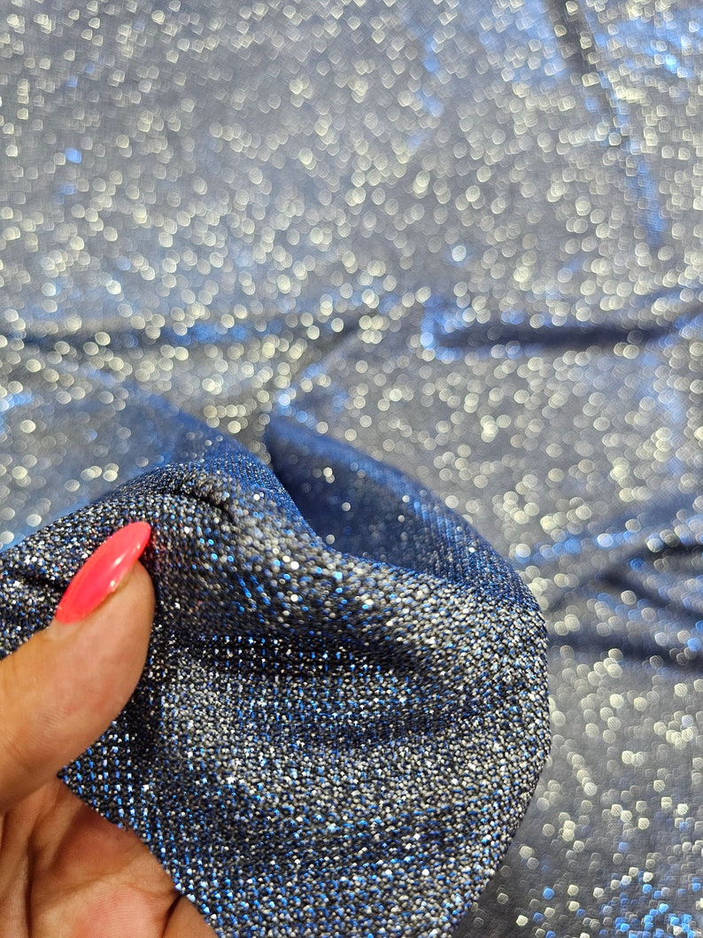 May include: Close-up of a silver sparkly fabric with a blue undertone. The fabric is being held by a hand with pink nail polish.