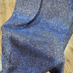 May include: A close-up of a dark blue fabric with a sparkly, glittery texture. The fabric has a fine, woven pattern and reflects light.
