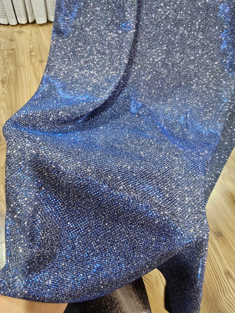 May include: A close-up of a dark blue fabric with a sparkly, glittery texture. The fabric has a fine, woven pattern and reflects light.