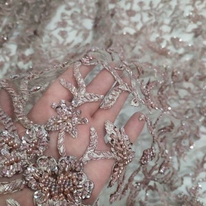May include: Close-up of a delicate, blush-coloured lace fabric with intricate floral embroidery and sparkling beads.