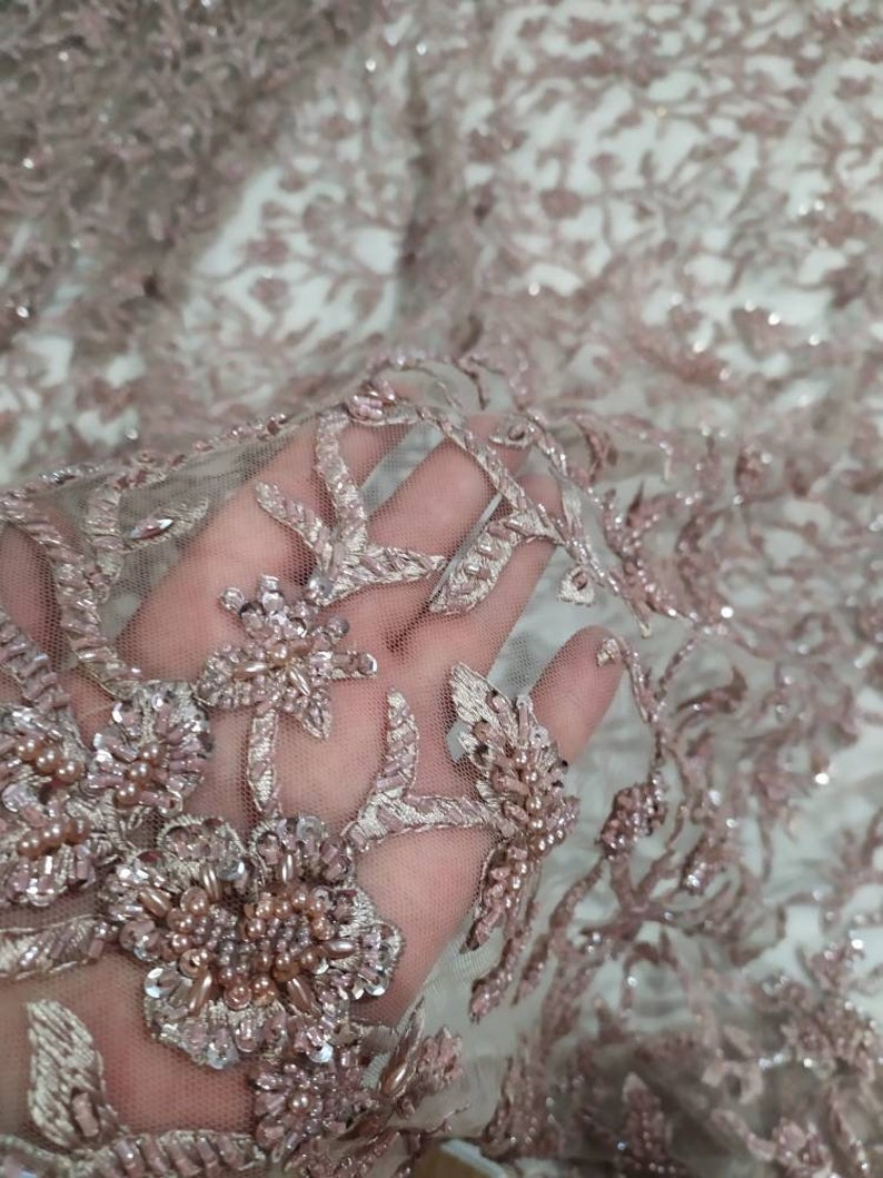 May include: Close-up of a delicate, blush-coloured lace fabric with intricate floral embroidery and sparkling beads.