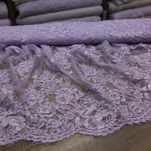 May include: A roll of lavender lace fabric with a floral pattern. The lace is draped over a surface, showcasing its intricate design and scalloped edge. The fabric is a soft, light purple colour.