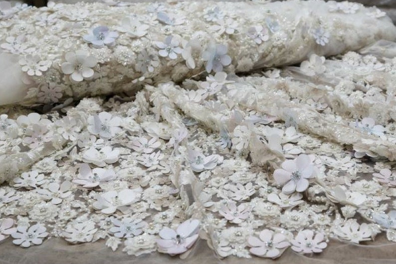 May include: White and light blue floral lace fabric with 3D flowers. The fabric has a delicate, sheer texture and is perfect for adding a touch of elegance to any project.