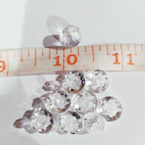 May include: A collection of small, clear, faceted glass beads arranged in a cluster. A white measuring tape is positioned across the beads, showing that each individual bead has a diameter of approximately 0.25 inches.