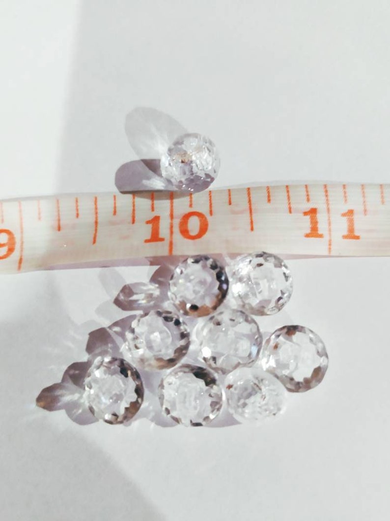 May include: A collection of small, clear, faceted glass beads arranged in a cluster. A white measuring tape is positioned across the beads, showing that each individual bead has a diameter of approximately 0.25 inches.