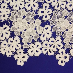 May include: White lace fabric with a delicate floral pattern. The lace is on a dark blue background.