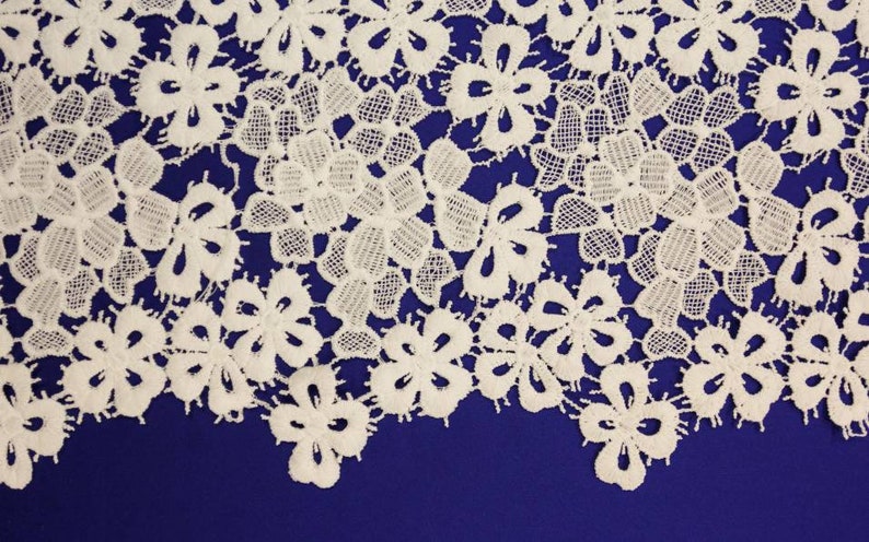 May include: White lace fabric with a delicate floral pattern. The lace is on a dark blue background.