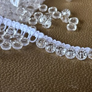 May include: A white button loop trim featuring a series of small, clear, round plastic buttons attached to a flexible fabric cord. The trim is arranged in a curved line, with additional loose clear buttons scattered around the main piece.