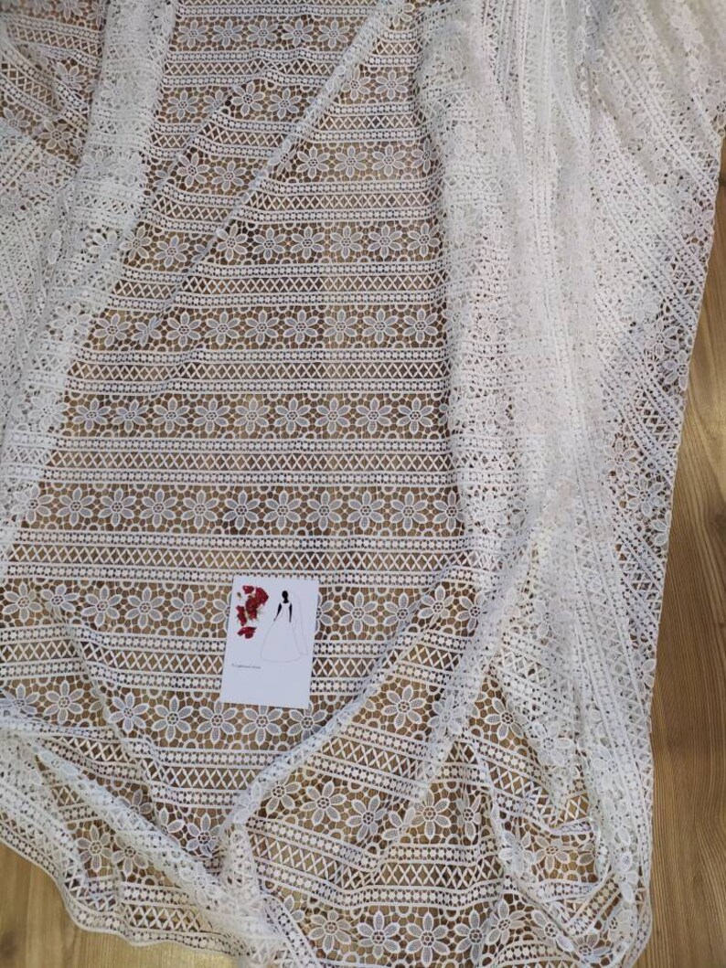 May include: White crochet fabric with a floral pattern. The fabric has a delicate, lacy texture and is perfect for crafting or sewing projects.