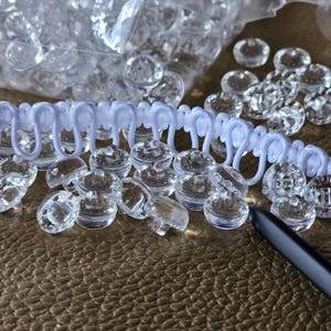 May include: A collection of small, clear, round plastic buttons with a shank back, accompanied by a length of white elastic loop trim. The buttons are scattered across the surface, with some contained in a clear plastic bag in the background.