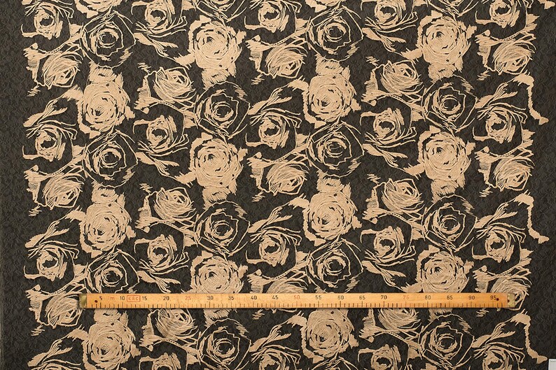 May include: Beige rose pattern on a dark brown fabric. The fabric is measured with a ruler in inches.