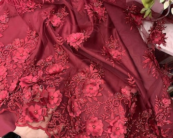 May include: Red lace fabric with 3D floral embellishments. The fabric has a sheer, mesh-like texture and features raised, burgundy-colored flowers.