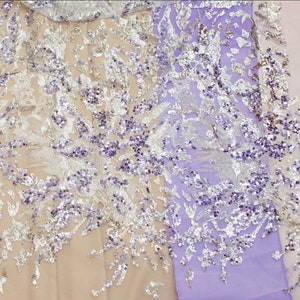 May include: A close-up of a sheer fabric with a floral pattern and silver and purple sequins. The fabric is a light purple color.