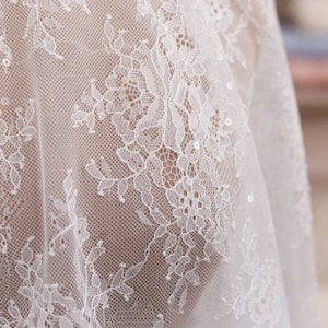May include: A sheer white lace fabric featuring an intricate floral pattern with delicate embroidery and small, scattered sequins that catch the light. The material has a fine mesh base, creating a semi-transparent texture with detailed botanical motifs.