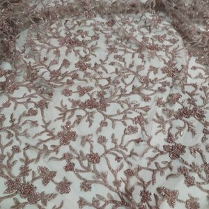 May include: A close-up of a delicate, rose gold beaded lace fabric with a floral pattern. The fabric is sheer and has a soft, shimmery finish.