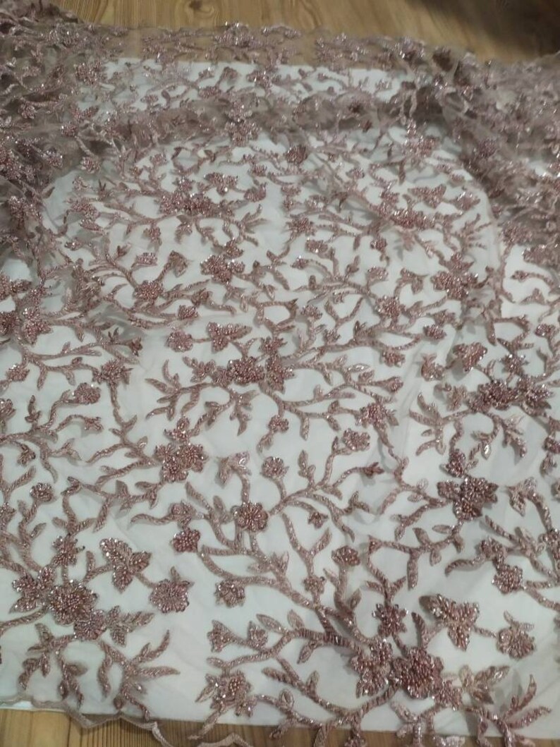 May include: A close-up of a delicate, rose gold beaded lace fabric with a floral pattern. The fabric is sheer and has a soft, shimmery finish.