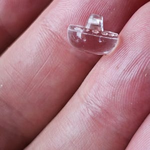May include: A small, clear, transparent button shaped like a boat. The button features a smooth, rounded hull design with a raised shank on the top center for attachment. The material is translucent and colorless, reflecting light on its surface.