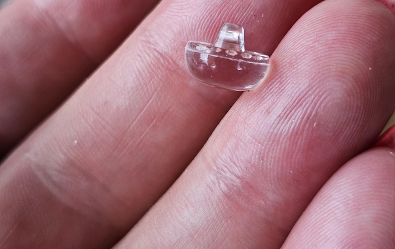May include: A small, clear, transparent button shaped like a boat. The button features a smooth, rounded hull design with a raised shank on the top center for attachment. The material is translucent and colorless, reflecting light on its surface.