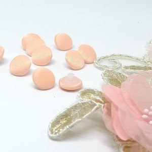 Peut inclure: Boutons recouverts de tissu de couleur pêche avec une fermeture à pression en plastique blanc. Les boutons sont disposés en demi-cercle sur une surface blanche.