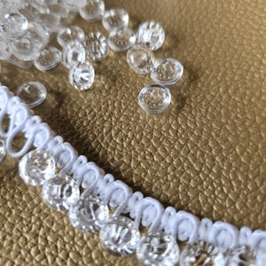 May include: A white loop trim featuring a series of clear, round, faceted buttons attached along the edge. Several additional loose, clear, round, faceted buttons are scattered nearby, showcasing their transparent, reflective, and circular design.