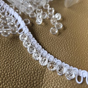 May include: A white button loop trim featuring a series of clear, circular, four-hole buttons attached to a flexible fabric tape. The trim is arranged in a curved line, with additional loose clear buttons scattered nearby on the surface.