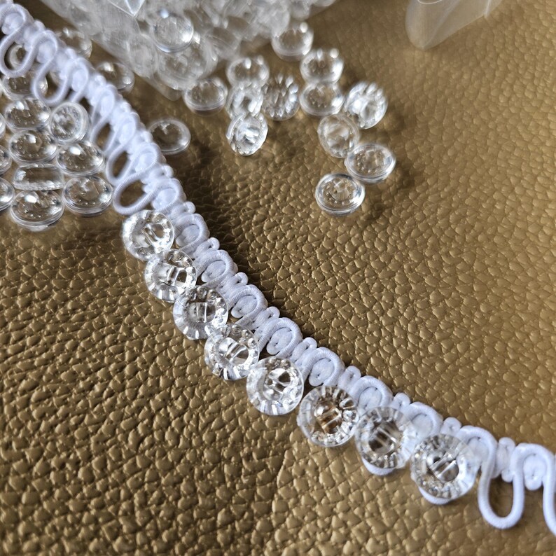 May include: A white button loop trim featuring a series of clear, circular, four-hole buttons attached to a flexible fabric tape. The trim is arranged in a curved line, with additional loose clear buttons scattered nearby on the surface.