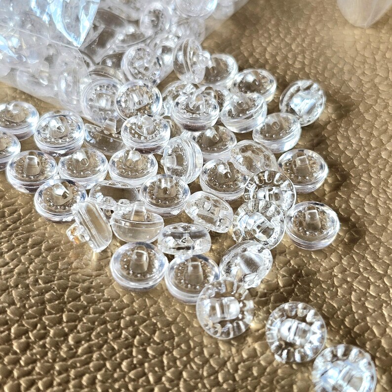 May include: A collection of small, round, clear plastic buttons with a smooth, transparent finish. Each button features a raised center with two holes for attachment, scattered across the surface in a loose, unorganized arrangement.