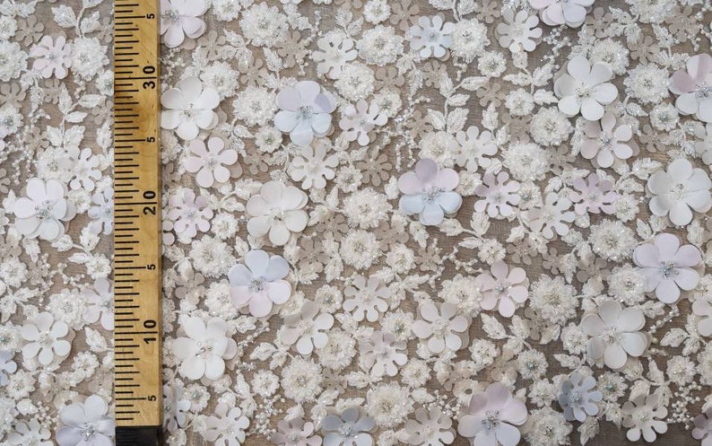 May include: White fabric with a delicate floral pattern. The fabric is covered in small, white flowers with a 3D effect. The flowers are arranged in a random pattern and are interspersed with small, white beads.