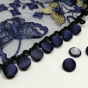 Puede incluir: Primer plano de botones cubiertos de tela azul marino con ribete negro y azul marino. Los botones están dispuestos en fila sobre una superficie blanca.
