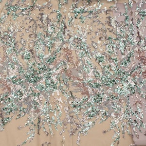 May include: A close-up of a fabric with a beige background and a pattern of silver, green, and pink sequins. The sequins are arranged in a swirling, abstract design.