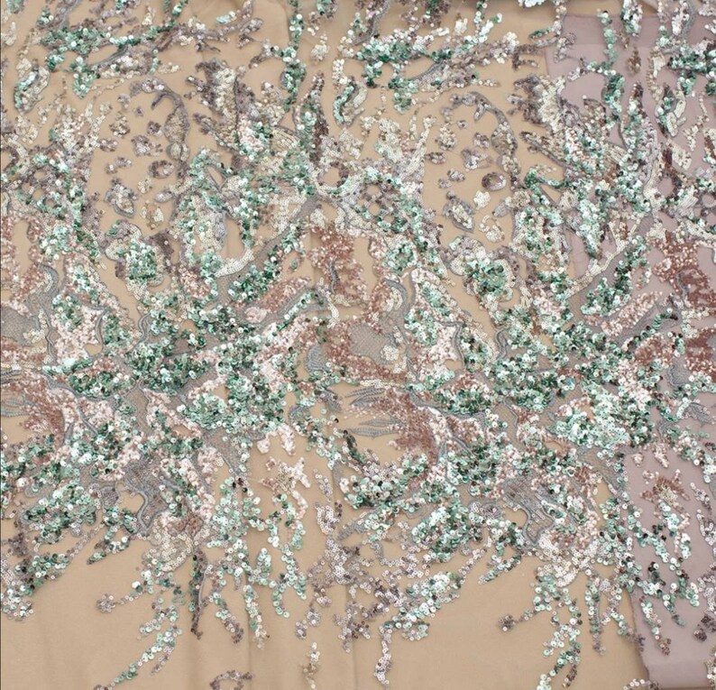 May include: A close-up of a fabric with a beige background and a pattern of silver, green, and pink sequins. The sequins are arranged in a swirling, abstract design.