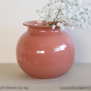 Puede incluir: Un jarrón redondo de cerámica de color coral con una abertura ancha, que contiene una ramita de flores blancas. El jarrón está colocado sobre una pila de libros, con el título "POST-IMPRESSIONISTS" visible. El jarrón tiene un acabado brillante.