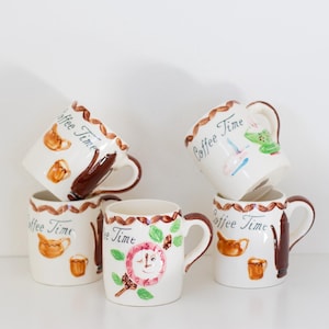 Könnte beinhalten: Eine Sammlung von fünf weißen Keramik-Kaffeetassen. Jede Tasse hat einen braunen Henkel und ist mit handgemalten Bildern von Kaffee-bezogenen Gegenständen und den Worten "Coffee Time" verziert. Die Tassen sind gestapelt und vor einem weißen Hintergrund angeordnet.