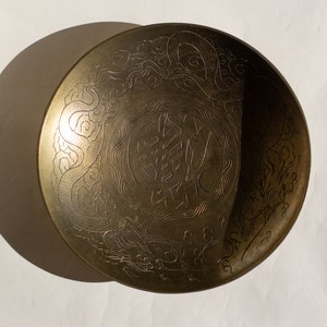 Op de afbeelding: Een ronde koperen schaal met een gegraveerd drakenmotief rondom een centraal karakter. Het metalen oppervlak heeft een warme, goudbruine kleur met gedetailleerde lijnen die het lichaam van de draak en wolkpatronen weergeven.