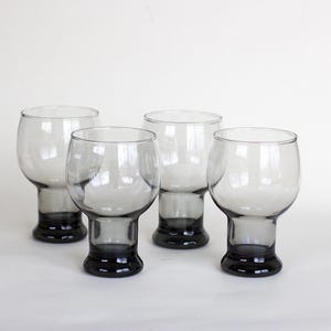 May include: Four clear glass goblets with a dark gray base. The goblets are arranged in a row on a white surface.