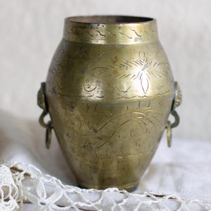 May include: A vintage brass vase with etched floral designs and small handles. The vase has a rounded body and a slightly flared rim. The brass has a warm, aged patina, suggesting it is an antique or vintage piece. The vase is displayed on a white cloth.